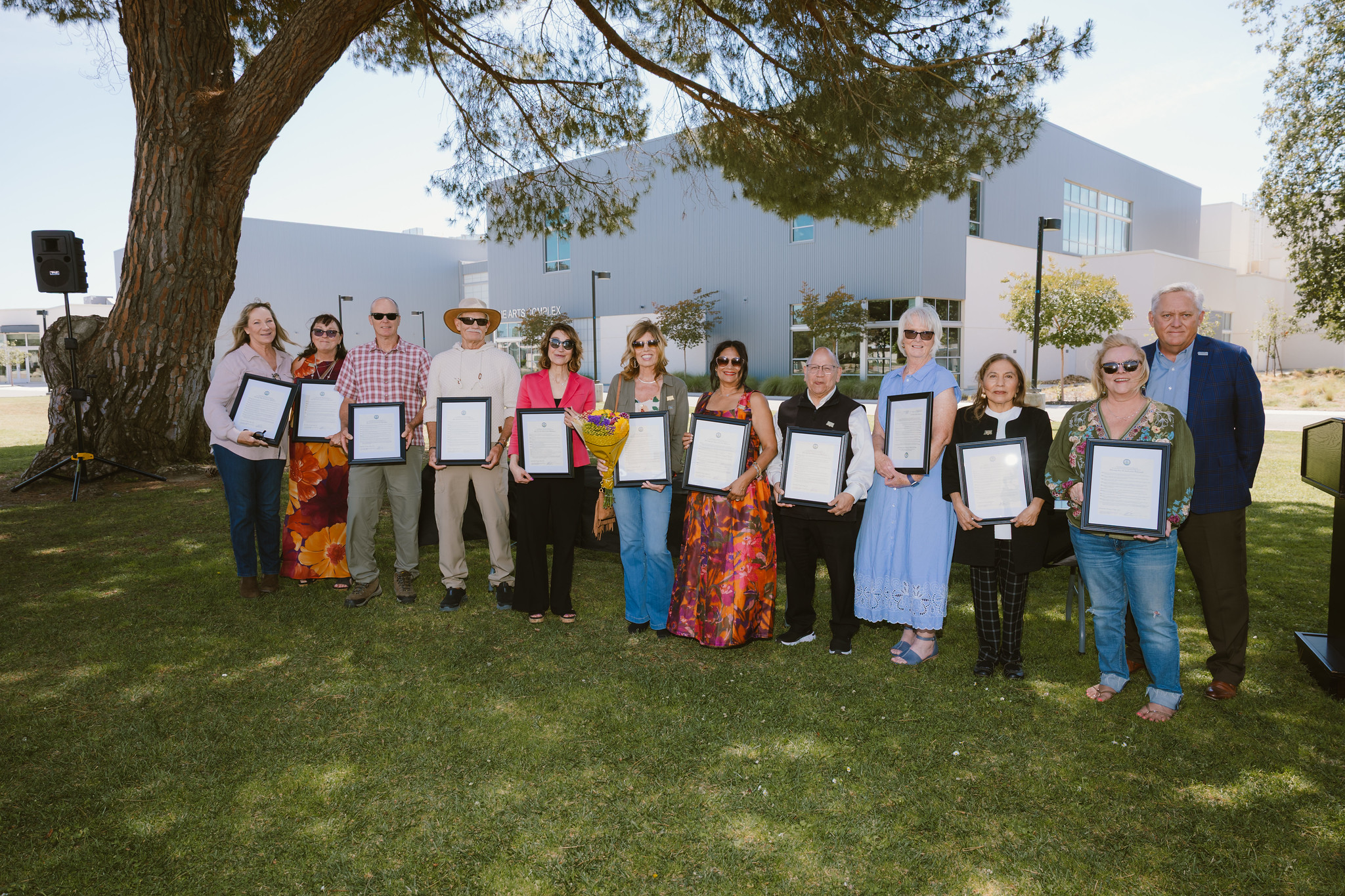 Retirees and honorees stand together outdoors during Allan Hancock College’s 2026 Retirements and Recognitions Ceremony. The group holds framed certificates while posing on a grassy campus area beneath a large tree, with the college’s Fine Arts Complex visible in the background.